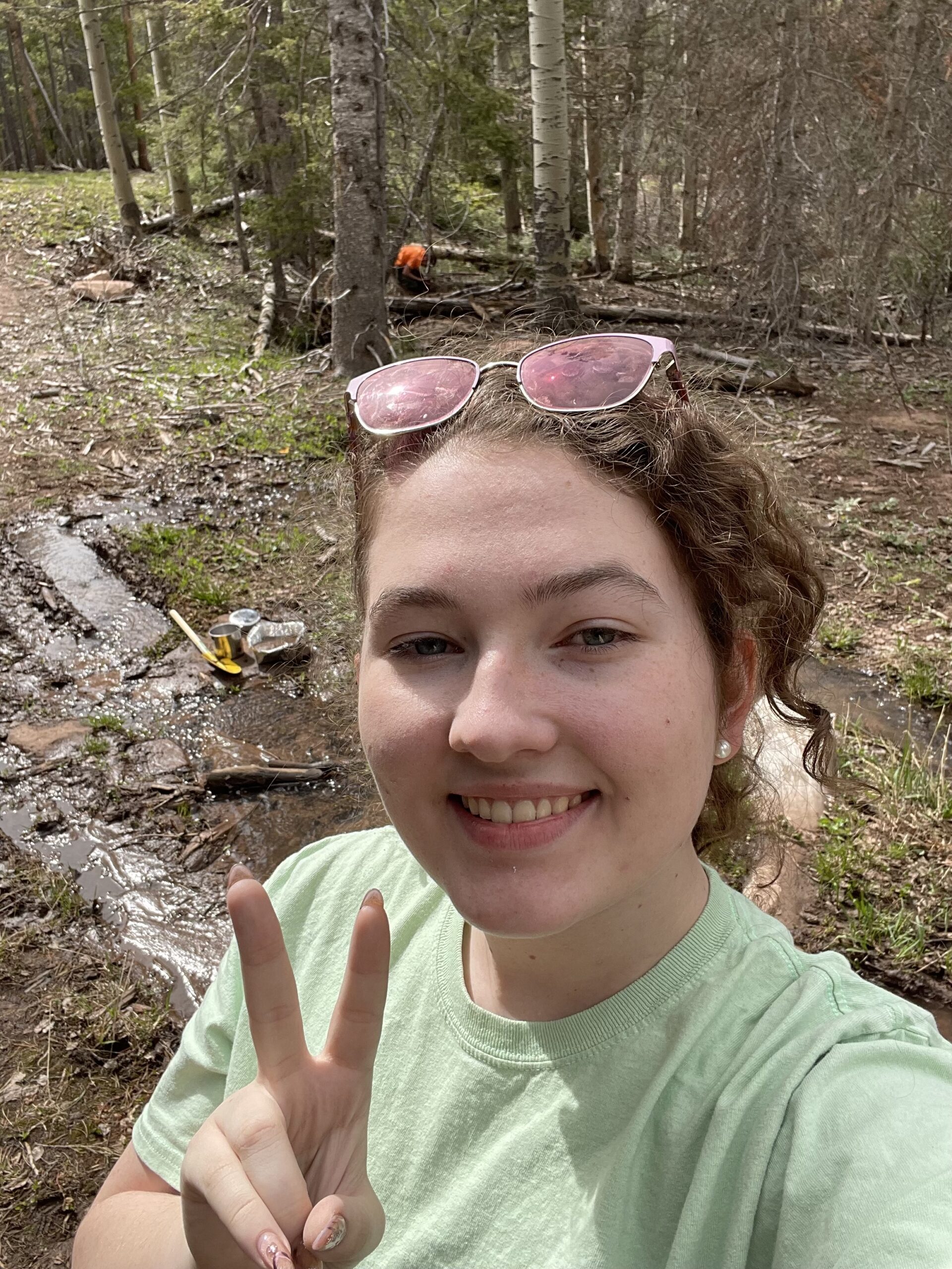 female student outside hiking wearing sunglasses on head and holding up a peace sign with her fingers.